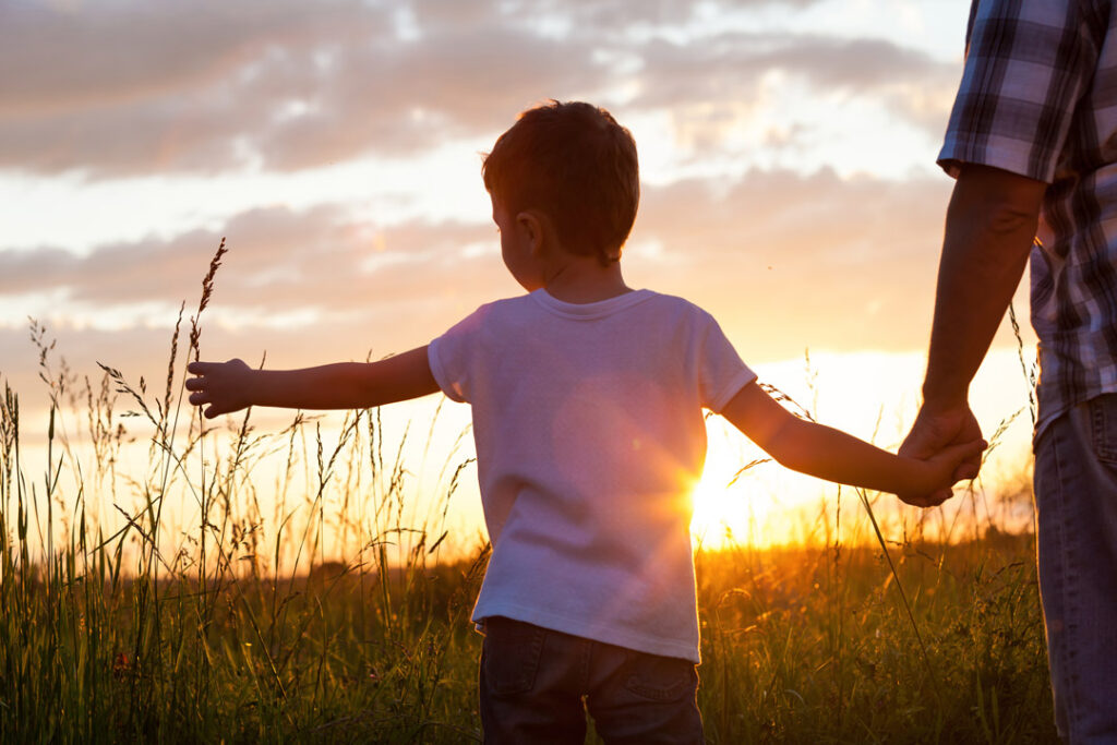 Young boy holds father's hand, who is out of frame, in a field at sunset